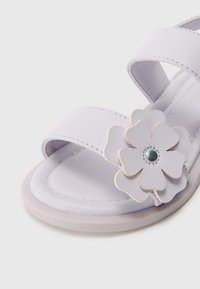 Lilla sandal med to brede stropper, med en blomsterdetalj med en lys blå midt. Glatt tekstur og polstret fotseng.