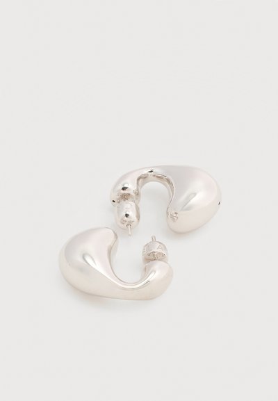 Ζευγάρι λαμπερών ασημένιων σκουλαρικιών καρφίτσας σε αφηρημένο σχήμα με στρογγυλεμένες, ακανόνιστες καμπύλες σε ανοιχτό φόντο.