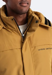 Senapsgul pufferjacka med dragkedja fram och svarta axelackcenter. Har en hög krage och mjuk textur. Logotyp synlig.