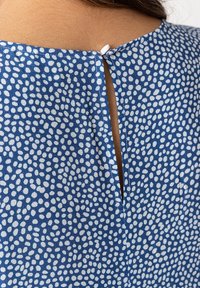 Blaues Kleid aus strukturiertem Stoff mit weißen Pünktchen. Der Rücken hat eine Schlüsselöffnungsdetails, die mit einem weißen Knopf befestigt ist.
