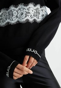 Maglione nero con un pannello in pizzo bianco con bordi frangiati sul petto e dettaglio del logo sui polsini. Tessuto liscio e forma aderente.