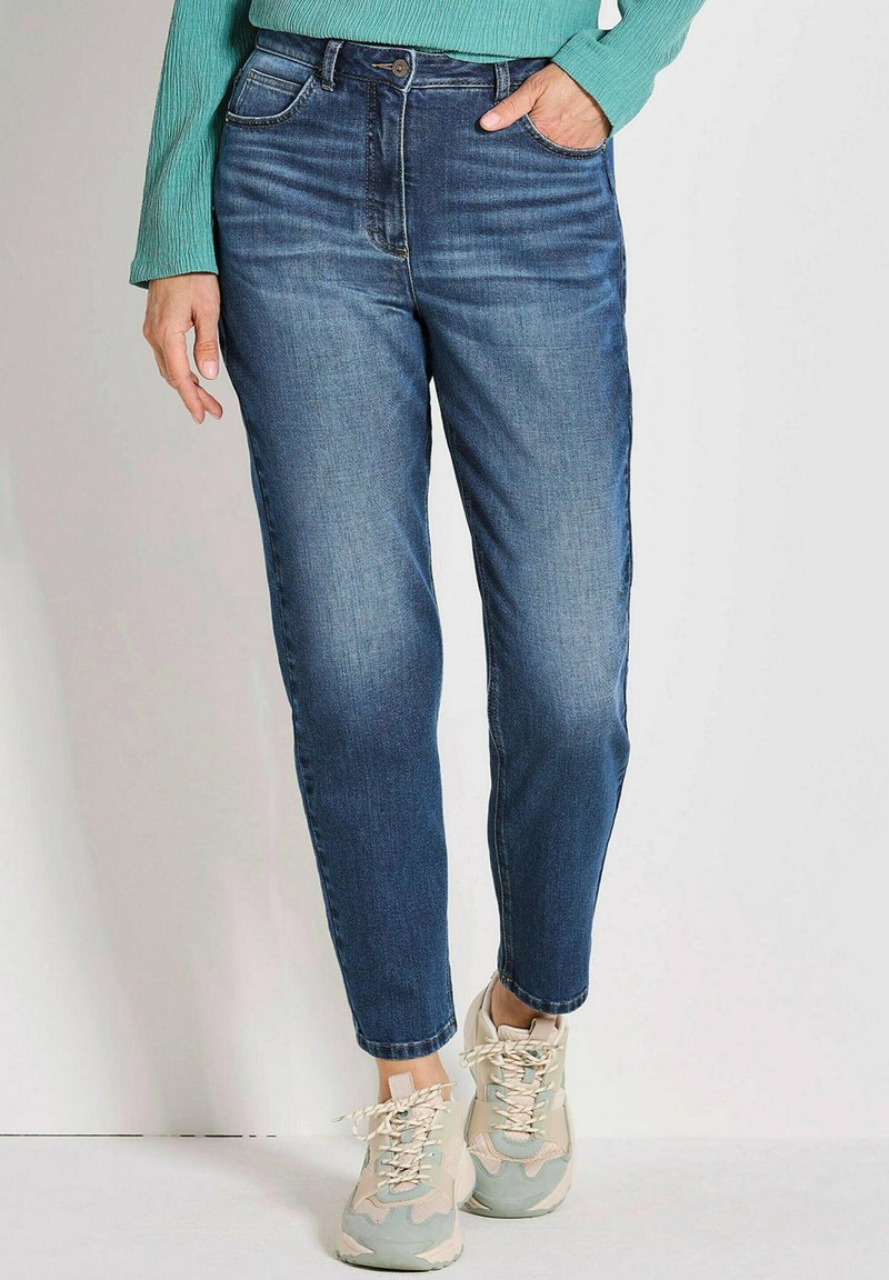 Jeans en denim taille haute de couleur bleu foncé avec une coupe droite, comportant des poches avant et une fermeture à boutons classique. Portés avec des baskets beiges.