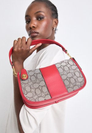 Vrouw met kort haar, gekleed in een wit topje, draagt een beige en rode designertas met monogram patroon over haar schouder.