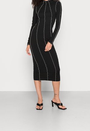 Femme portant une robe midi noire à manches longues avec des coutures verticales blanches ondulées et des sandales noires à talons ouvertes, sur un fond uni.