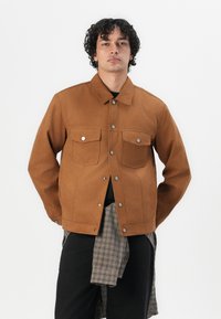 Bruine denimjas met een knoopsluiting aan de voorkant, twee borstzakken en een rechte snit. Voorzien van metalen drukknoopjes en een gladde textuur.