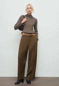 Donna con capelli biondi che indossa un maglione di maglia marrone, pantaloni larghi marroni con una cintura beige, stivali neri alla caviglia e tiene in mano una borsa marrone.