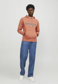 Sudadera de color naranja claro con cordones, bolsillo delantero y logo. Combinada con unos pantalones vaqueros azules holgados y zapatillas blancas. Diseño simple y casual.