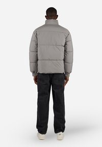 Pufferjakke i lys grå, med høy krage og horisontalt quiltet mønster. Kombinert med svarte, løstsittende jeans og hvite joggesko.