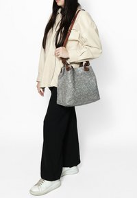 Borsa grigia con texture e stampa floreale, manici in pelle marrone, forma a secchiello. La modella indossa una camicia oversize alla moda e pantaloni neri a gamba larga.