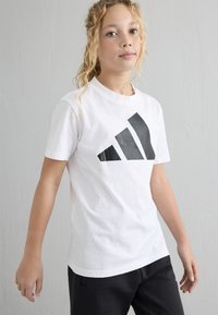 Balta kokvilnas T-krekls ar lielu melnu trīsrindas logotipu uz priekšpuses. Īsās piedurknes un apaļš kakla izgriezums.