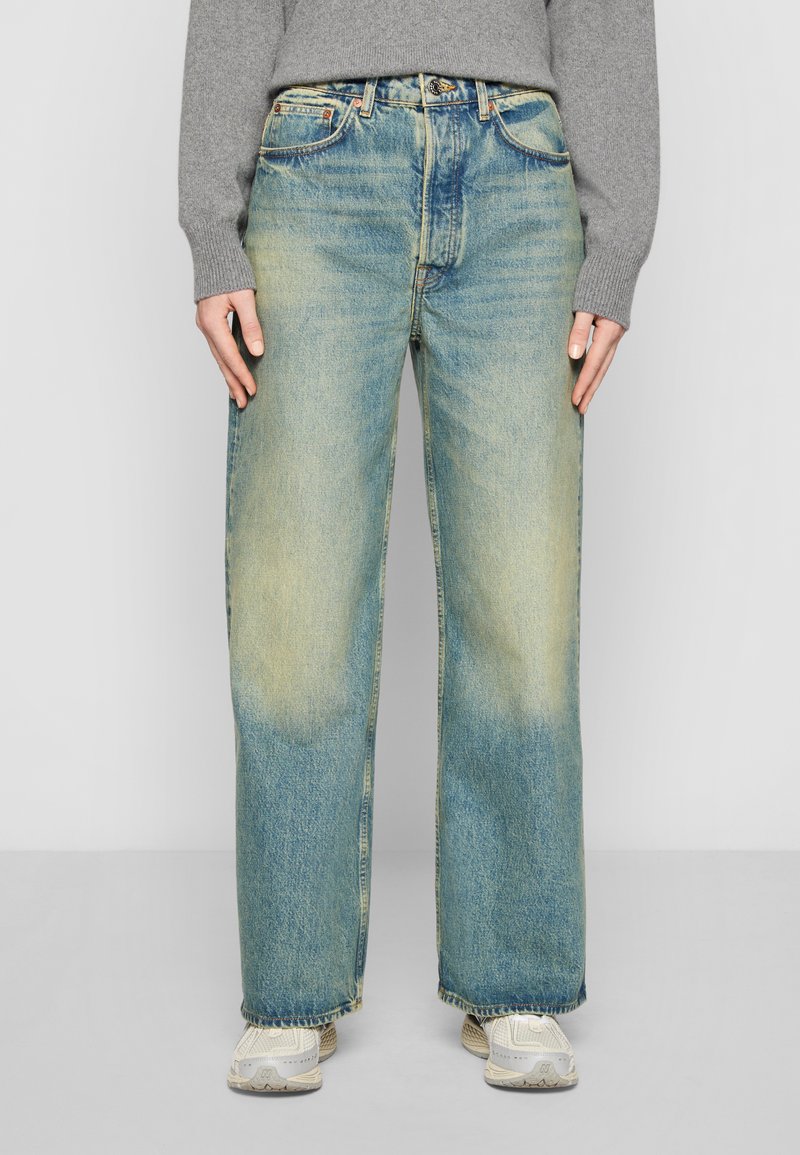 Jean large bleu clair avec un délavage, présentant un design classique à cinq poches et des détails en métal, associés à des baskets neutres.