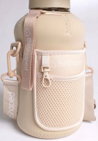 Bouteille d'eau BeShaped avec une housse en néoprène beige, une poche en maille, une sangle ajustable et du matériel beige, avec des coutures visibles.