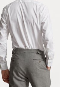 Mies, joka pitää yllään valkoista pitkähihasta paitaa, joka on sisätetty harmaisiin juhlahousuihin, joissa on napilliset takataskut ja sivusäätimet, ja yksi käsi on taskussa.