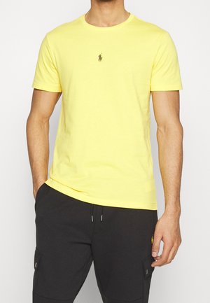 Mann trägt ein schlichtes gelbes Kurzarm-T-Shirt mit kleinem Logo auf der Brust und schwarze Hose mit Seitentaschen.