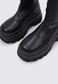 Botas de tobillo de cuero negro con una plataforma gruesa y suela de goma texturizada, con un acabado liso en la parte superior y detalles de costura.