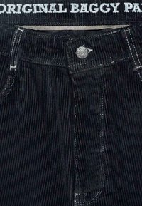 Crne široke hlače od kordura s vertikalnim rebrima, bijelim šavovima, metalnim gumbom i pojasom na kojem stoji "ORIGINAL BAGGY PANTS" u bijelom tekstu.