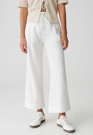 Femme portant un pantalon blanc à jambes larges avec ourlets retroussés, un haut court beige noué devant et des baskets blanches avec des rayures grises.
