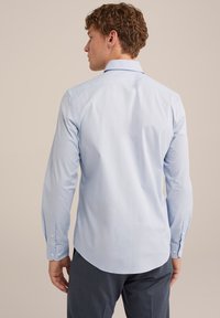 Chemise à manches longues bleu clair avec col boutonné, texture lisse et ourlet incurvé, associée à un pantalon gris foncé.