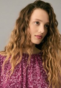 Haut violet à motif floral avec un col smocké et une coupe décontractée, associé à des cheveux lâches et ondulés aux tons naturels.