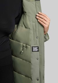 Geacă puffer de culoare verde măslinie, cu un design quilted, etichetă de marcă vizibilă și nasturi tip snap. Interiorul are un căptușeală moale și izolantă.