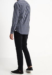 Uomo che indossa una camicia a scacchi blu e nera, pantaloni neri e sneakers nere, in piedi con le spalle rivolte.