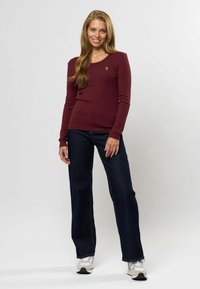 Top ribbed de culoare bordeaux cu mânecă lungă și guler rotund, asortat cu blugi cu croială largă de culoare albastru închis. Modelul poartă teniși gri, stând pe un fundal deschis.