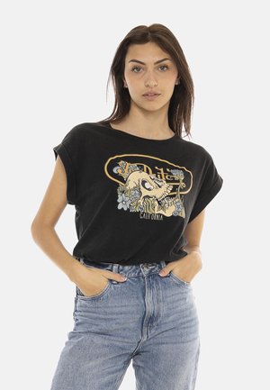 Jeune femme aux longs cheveux foncés portant un t-shirt graphique noir avec un motif de crâne et de fleurs et un jean bleu clair taille haute.