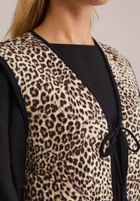 Vesta s leopardím potiskem vyrobená z měkké texturované látky, s černým lemem a předním zavazováním, nošená přes černé tričko.