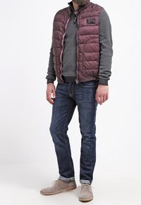Gilet trapuntato bordeaux sopra un maglione grigio testurizzato, abbinato a jeans blu scuri e scarpe in suede beige con lacci e suole chiare.