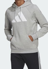 Sudadera gris hecha de tejido suave, con un gran logo blanco en el pecho, bolsillo frontal tipo canguro y capucha con cordón.