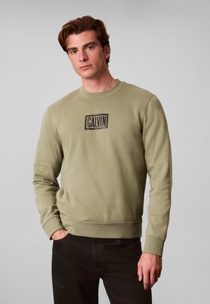 Giovane uomo che indossa una felpa verde oliva chiaro con il logo "CALVIN" sul petto, abbinata a pantaloni neri, in piedi davanti a uno sfondo grigio uniforme.