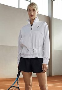 Jauna sieviete ar tenisa raketi, tērpusies baltā rāvējslēdzēja jaciņā un melnos šortos, stāvot iekštelpu tenisa kortā.