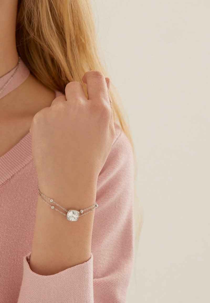Femme portant un bracelet double-chaîne en argent avec une grosse pierre précieuse carrée claire, vêtue d'un haut à manches longues rose poudré.