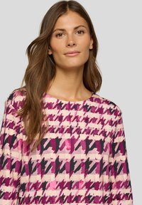 Frau mit langen braunen Haaren, die ein Oberteil mit abstraktem Hahnentrittmuster in Pink, Lila und Schwarz trägt, blickt nach vorne mit neutralem Ausdruck.