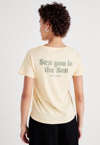 Světlý žlutý bavlněný tričko s uvoln�ěným střihem. Na zadní straně je zelený text s nápisem "Sea you in the Sun" a "PEPE JEANS."