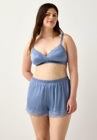 Una donna con lunghi capelli scuri posa con sicurezza, indossando un reggiseno blu con ricami in pizzo e pantaloni coordinati su uno sfondo neutro.