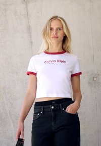 White cropped t-shirt with red trim and embossed "Calvin Klein" text, paired with high-waisted black jeans and a black accessory.