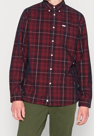 Homme portant une chemise rouge et noire à carreaux à manches longues boutonnée et un pantalon vert olive, debout avec les bras détendus.