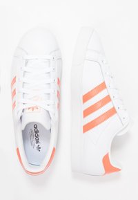 Vit Adidas-sneakers med korallfärgade tre ränder, snörning och hälaccenter, visade från ovan och sidan på en vit bakgrund.