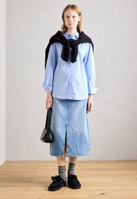 Femme portant une chemise bleu clair à boutons, jupe en denim mi-longue avec fente sur le devant, pull noir noué sur les épaules, tenant un sac à main noir.
