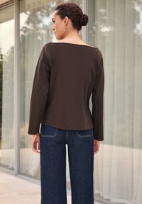 Maglia a maniche lunghe marrone con scollo quadrato, abbinata a jeans ampi a gamba larga di colore blu scuro. Il tessuto sembra liscio, con semplici dettagli di cucitura.