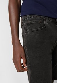 Jean en denim noir avec une coupe slim et une légère décoloration. Le tissu a une texture douce, et le design comprend des poches avant et des surpiqûres contrastantes.