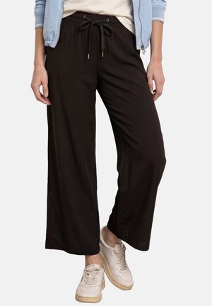 Persona che indossa pantaloni neri larghi con coulisse, giacca azzurro chiaro, camicia bianca e scarpe da ginnastica bianche, in piedi davanti a uno sfondo semplice.