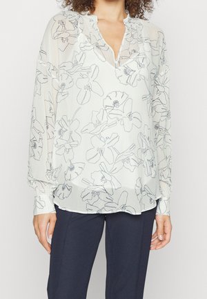 Vrouw draagt een witte blouse met lange mouwen met zwart bloemmotief en donkerblauwe broek, staand tegen een effen achtergrond.