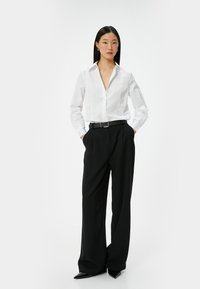 Blusa blanca de manga larga con cierre de botones combinada con pantalones de pierna ancha negros de talle alto y un cinturón negro; un aspecto elegante y ajustado.