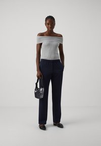 Grijze ribgebreide top met blote schouders, marineblauwe ontspannen pasvorm broek, zwarte schoenen, in de hand een kleine zwarte met pailletten versierde handtas, tegen een effen achtergrond.