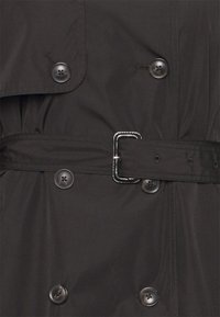 Tummanruskea trench-coat, jossa on kaksirivinen malli, suuret mustat napit ja yhdisteessä oleva vyö, jossa on suorakulmainen metallisolki.