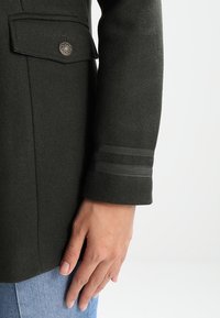 Tmavě zelený vlněný kabát s přední kapsou a detailem na knoflík, s dvěma vodorovnými pruhy na manžetě. Hladká textura.