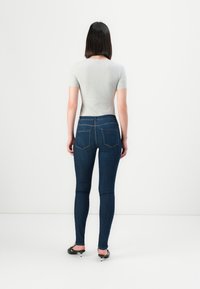 Woman with straight dark hair wearing a grey fitted short-sleeve top, dark blue skinny jeans, and black heels, standing facing away.