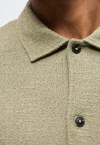 Κοντινό πλάνο υφασμάτινου ανοιχτό ελιάς πράσινου πόλο με μαύρα κουμπιά που φοριέται από άτομο με μέτριο καφέ τόνο δέρματος.